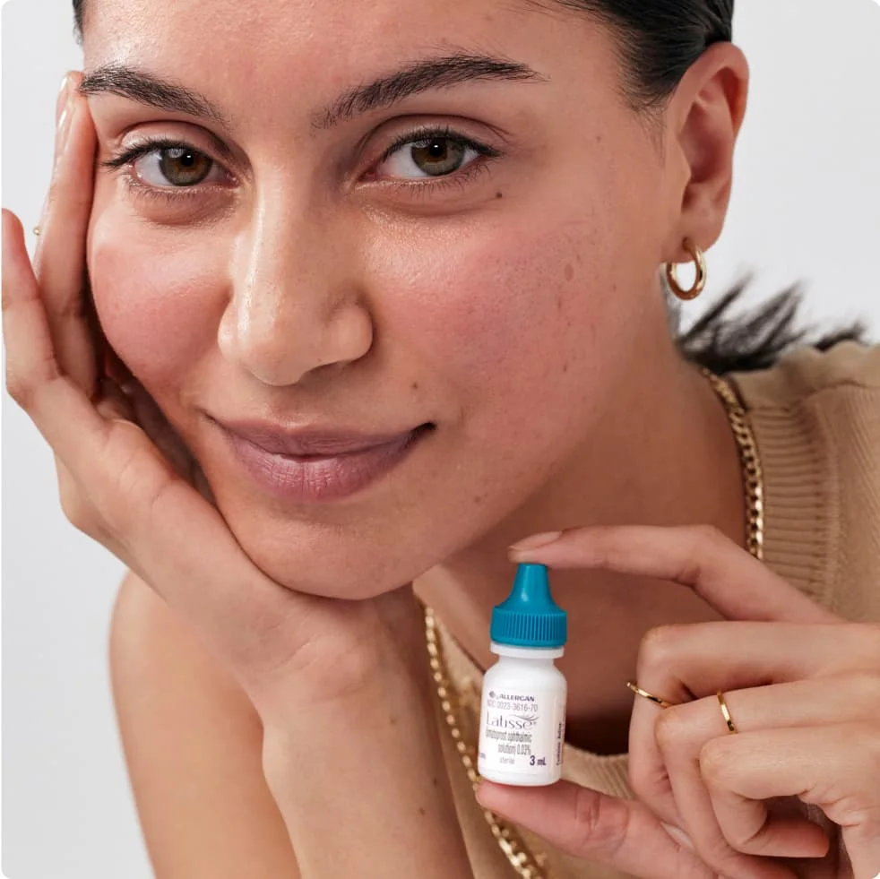 Woman with her hair pulled back leans her chin into her palm while holding a latisse bottle in the other